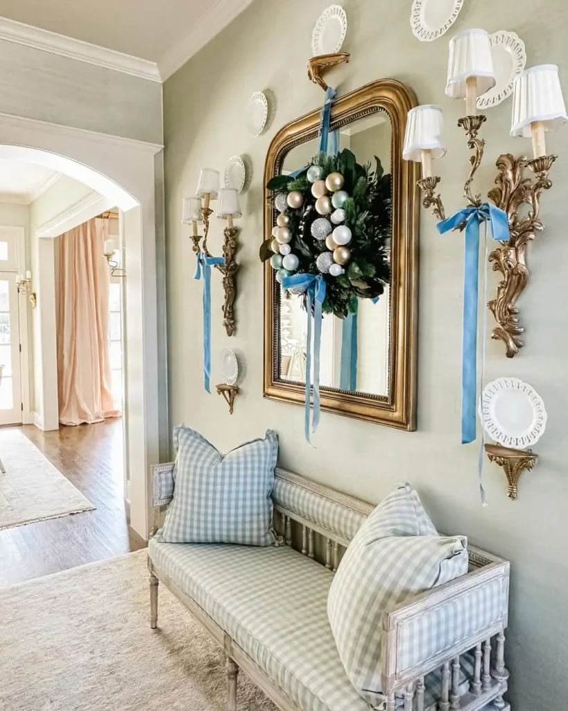 A hallway with a gold-framed mirror, wall sconces, and decorative plates above a gingham-patterned bench. DIY entryway Christmas decorations, like a wreath with ornaments and blue ribbons, hang on the mirror.