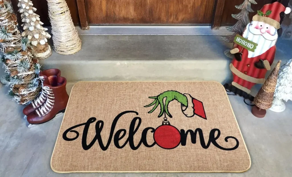 A doormat with "Welcome" written on it, featuring a green hand in a Santa sleeve holding a red ornament, placed at a festive porch with Christmas decorations.