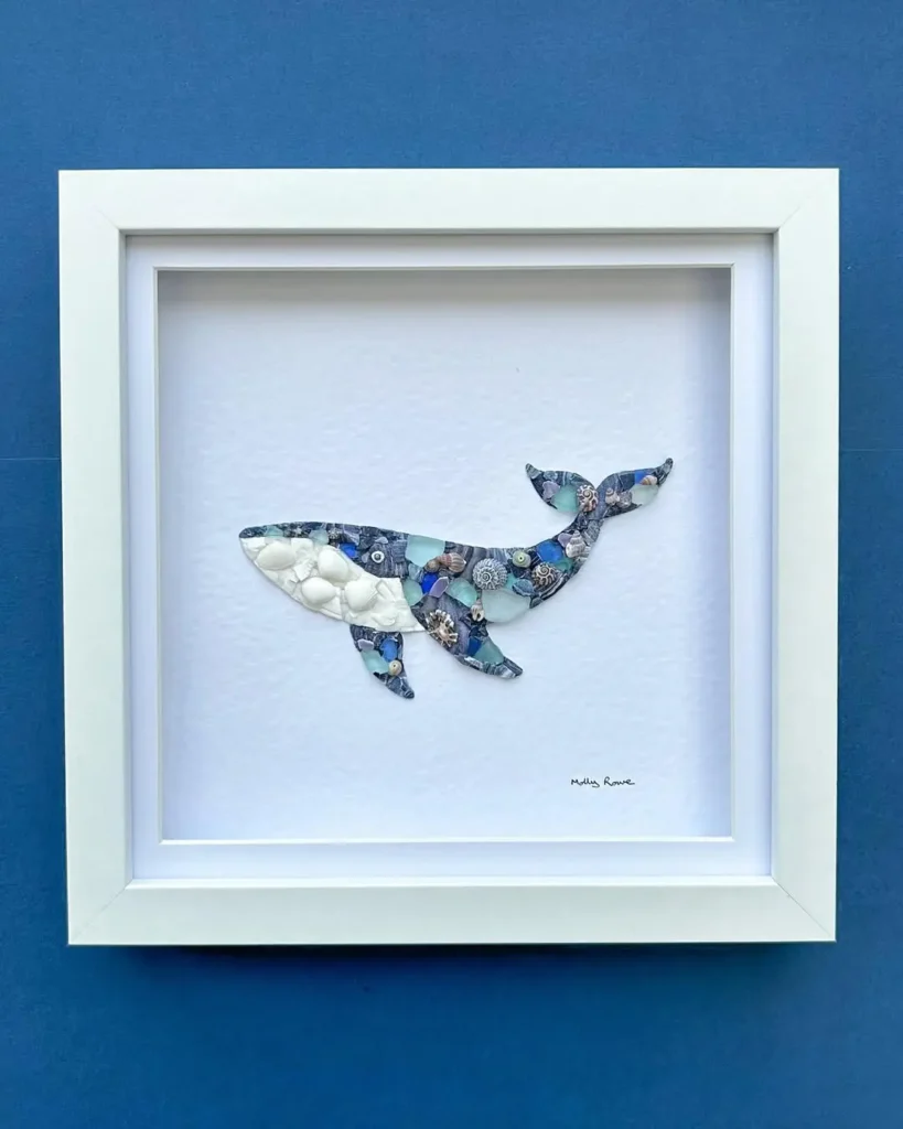 Framed artwork of a whale crafted from assorted blue and white patterned seashells, displayed against a white background with a blue outer wall.