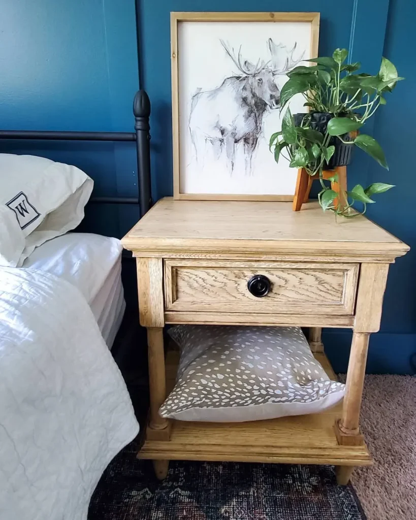 A reclaimed wooden nightstand with a drawer and lower shelf holds a pillow, framed moose sketch, and potted plant, beside a bed with white linens against a blue wall.