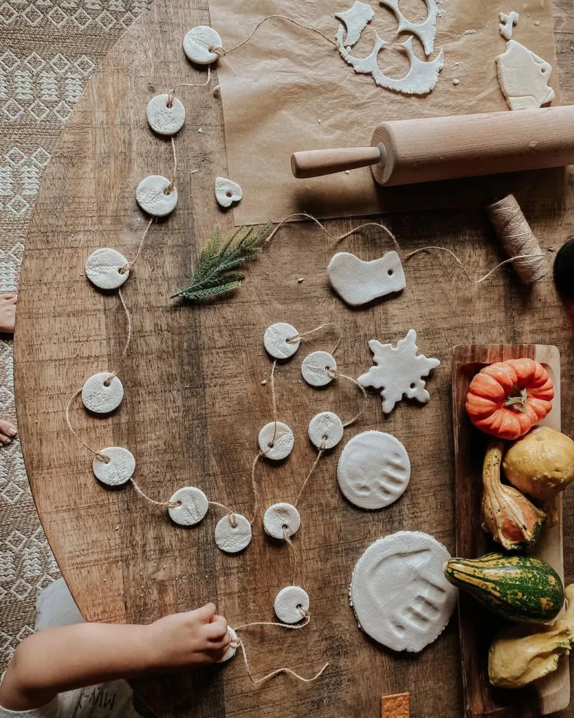 A child’s hand arranges salt-dough Christmas cookie ornaments on a wooden table with twine, a rolling pin, parchment paper, and assorted gourds.