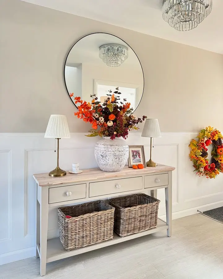 A console table with two wicker baskets, a large vase of autumn flowers, two lamps, and a framed photo sits below a round mirror in a bright entryway, creating inviting Thanksgiving entryway decor with natural elements.