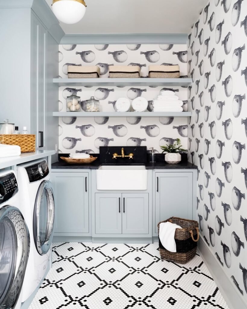 A modern laundry room with light blue cabinets, open shelves holding towels and jars, a black countertop, farmhouse sink, and black and white wallpaper design for a bold modern laundry room. Washer and dryer on the left.