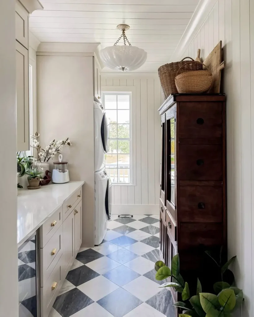 A narrow laundry room with black and white checkered floor, stacked washer and dryer, white cabinets, wooden hutch, baskets, plants, and a window letting in natural light—use easy to clean and maintain surfaces to avoid a common laundry design mistake.
