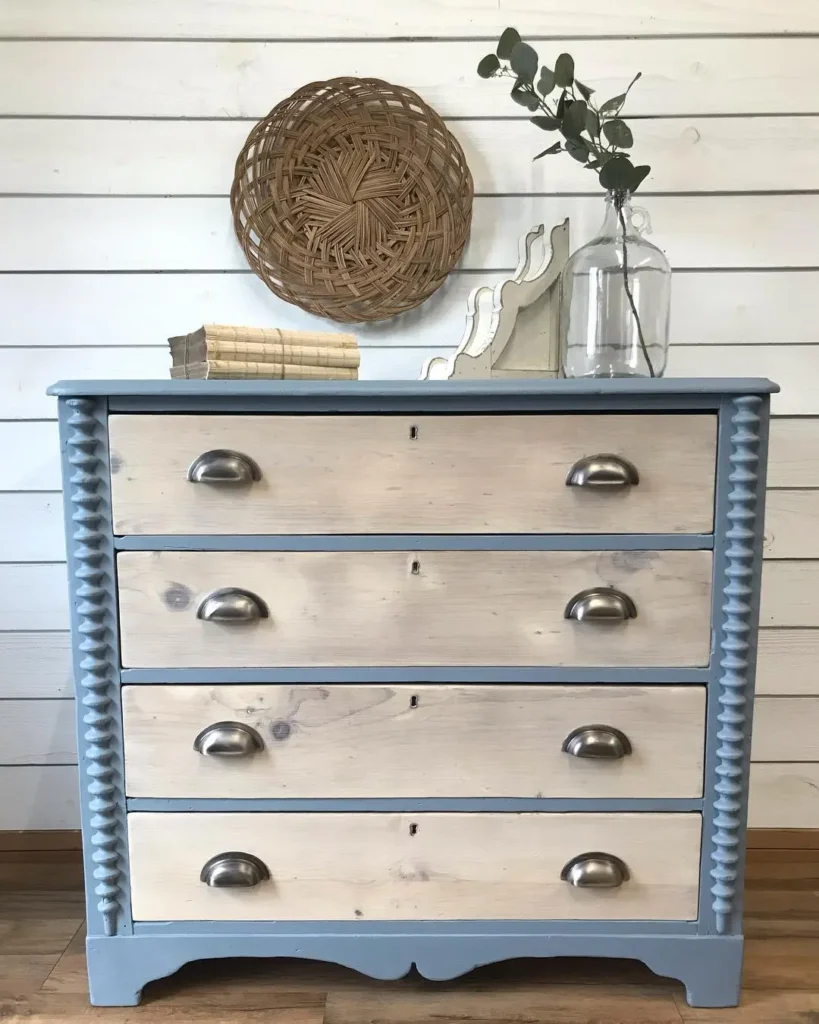 An affordable rustic bedroom furniture piece, this pine dresser features four drawers, a blue frame, and natural wood fronts. On top are books, a wicker tray, a glass vase with leaves, and two white decorative objects.