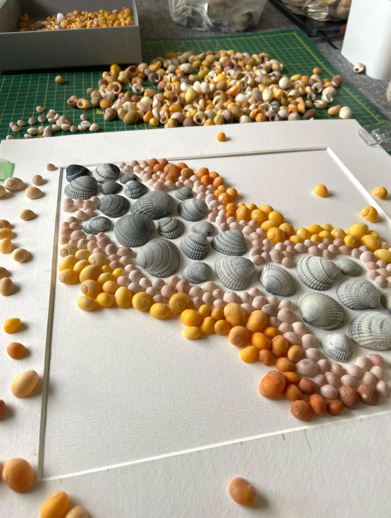 A white canvas partially covered with small and medium seashells, arranged in a gradient pattern, with more loose shells scattered on the table.