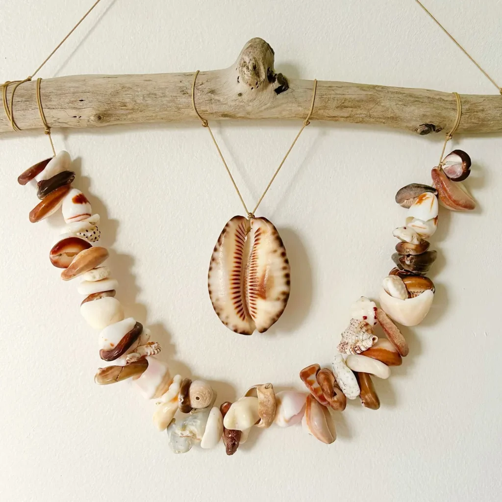 A decorative wall hanging made of driftwood, seashells, and a large cowrie shell, suspended with string against a plain white wall.