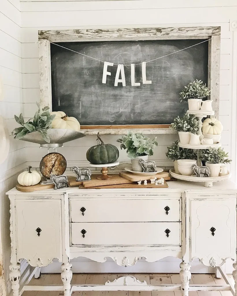 A vintage white sideboard is decorated with small pumpkins, greenery, stacked bowls, and farmhouse-style animal figurines for charming farmhouse fall decor; a chalkboard above displays the word "FALL.