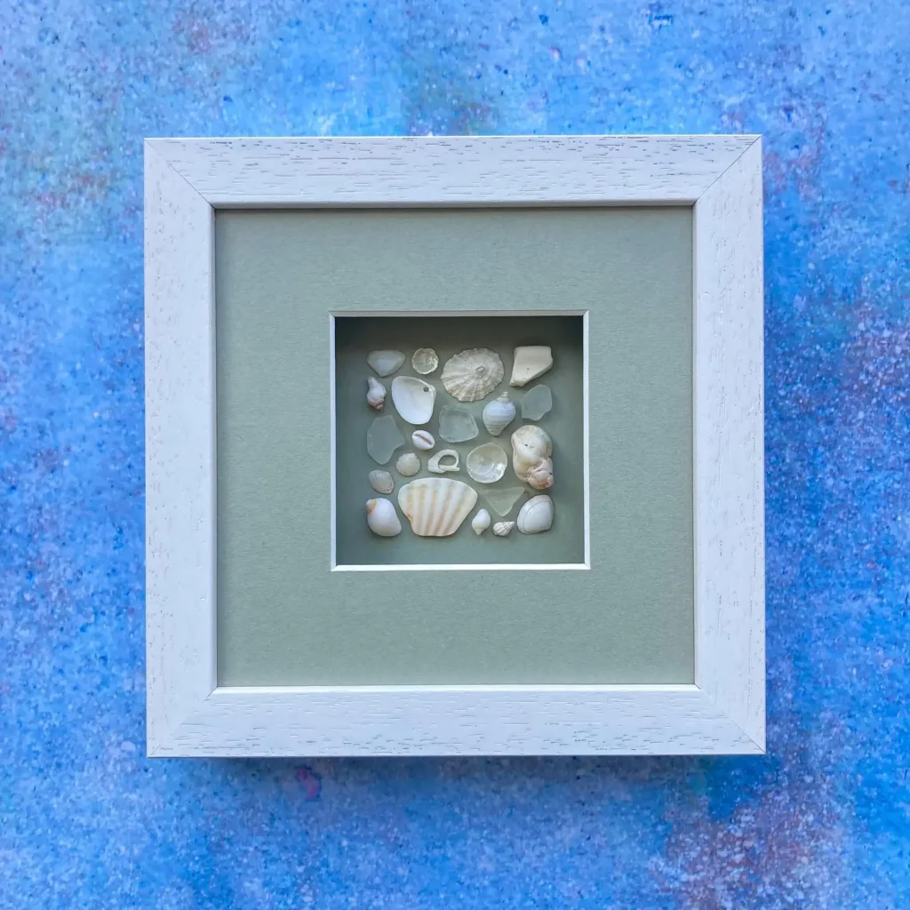 A white-framed, matted display of assorted small seashells arranged in a square, set against a textured blue background.