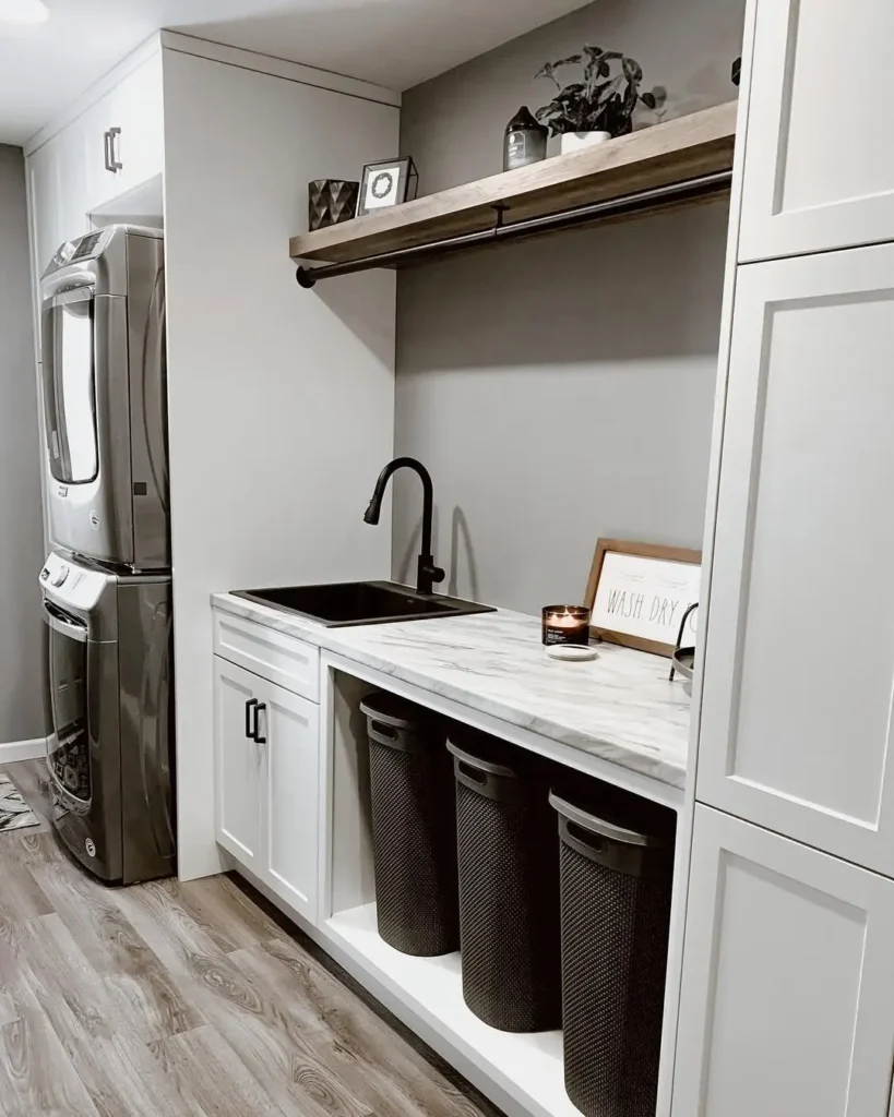 Modern laundry room with stacked washer and dryer, black sink, marble countertop, three black laundry baskets for a sorting system to avoid common laundry mistakes, and a shelf with decor items above.