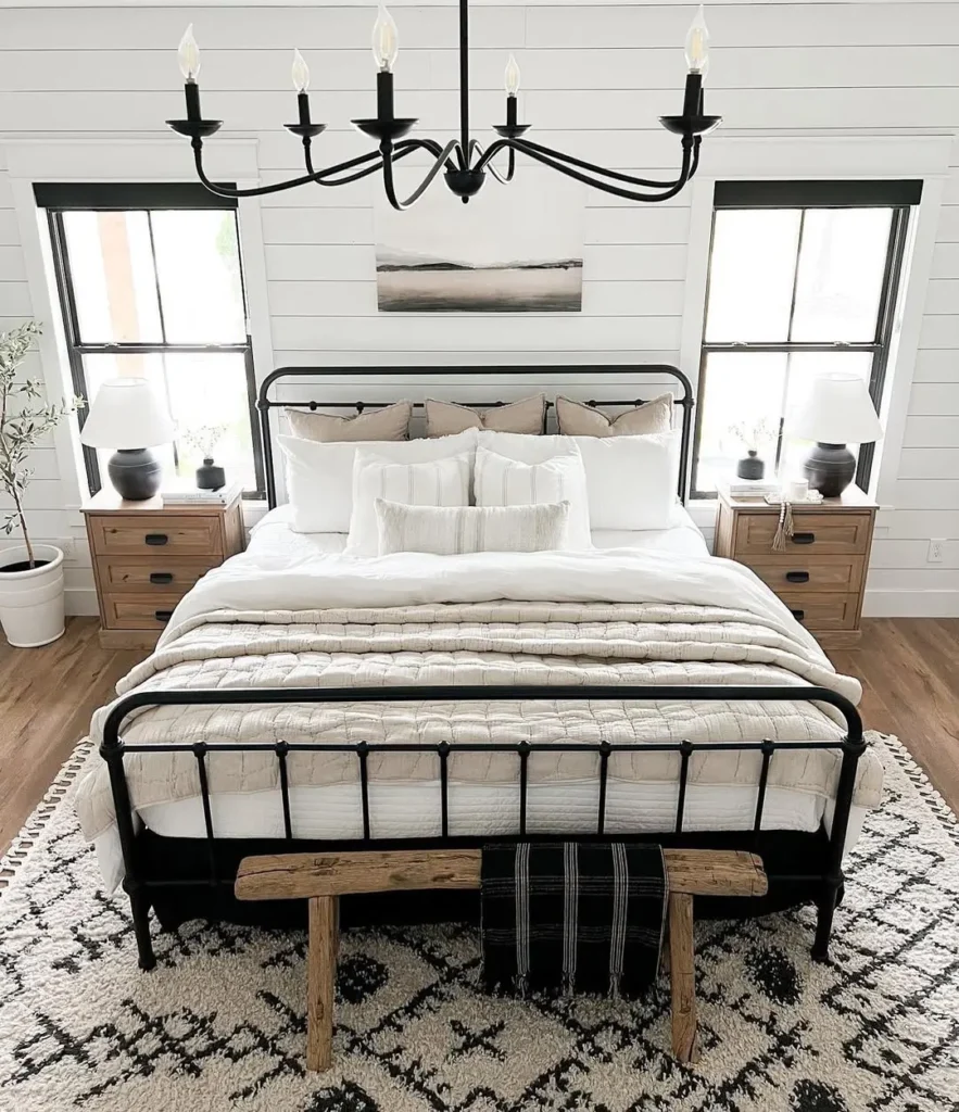 A neatly made bed with white bedding, black metal frame, and wooden bench sits between two windows; matching nightstands with lamps are on each side, showcasing rustic bedroom furniture beneath a striking black chandelier.