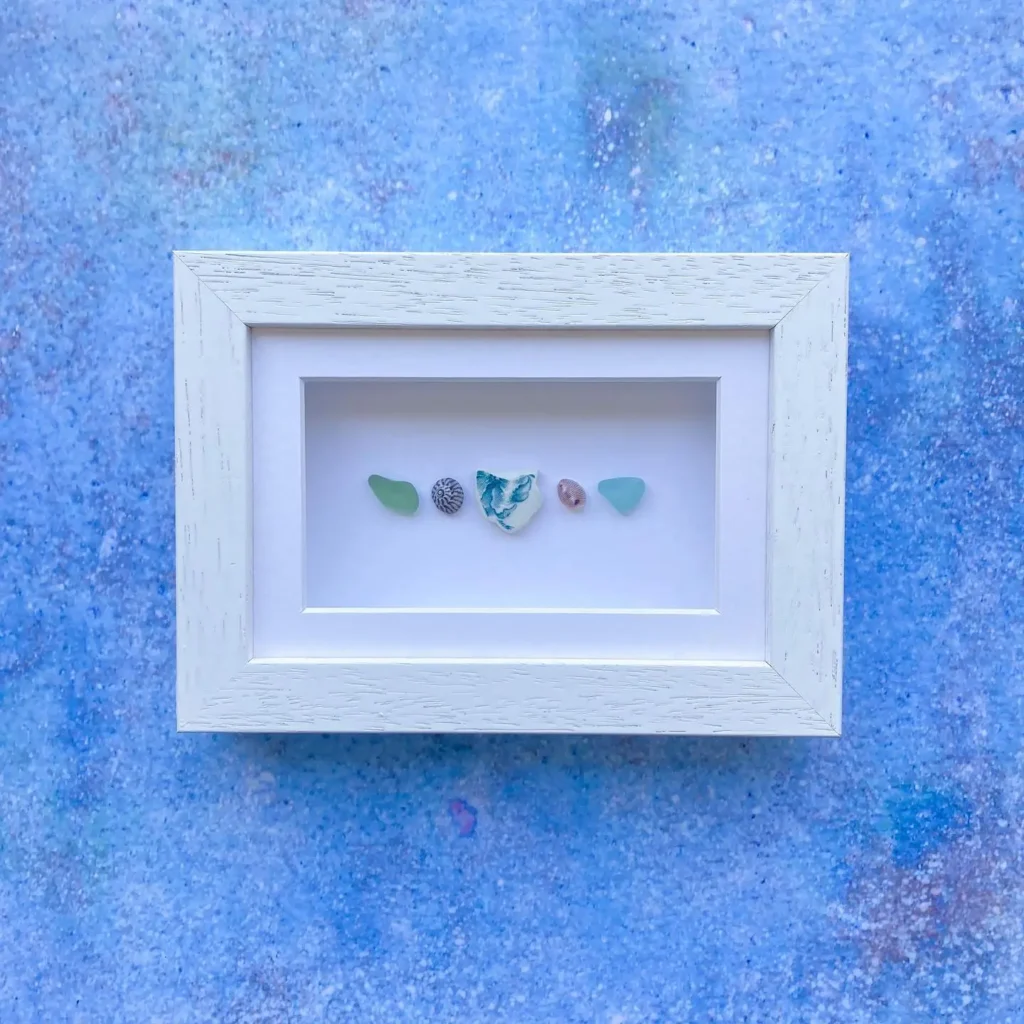 A white frame displays five small, colorful sea glass and ceramic pieces arranged in a row on a white background, set against a blue textured surface.