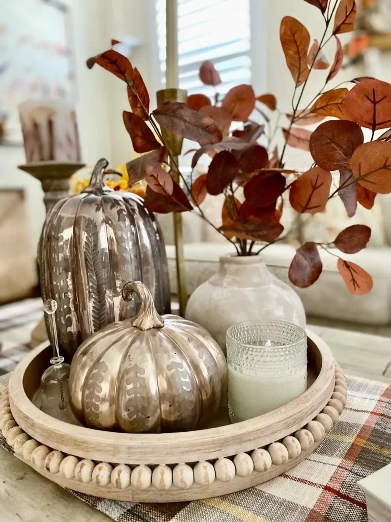 A wooden tray creates a modern fall centerpiece with two metallic pumpkins, a small glass stirrer, a white vase filled with brown leaves, and a lit white candle on a plaid tablecloth.