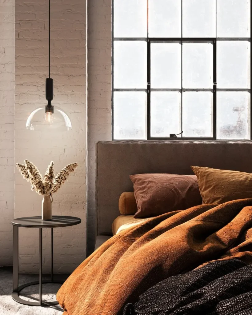 A modern bedroom with a large window, brown bedding, a beige headboard, a round side table, and modern fall textured decor like a vase with dried flowers and a hanging glass pendant light.
