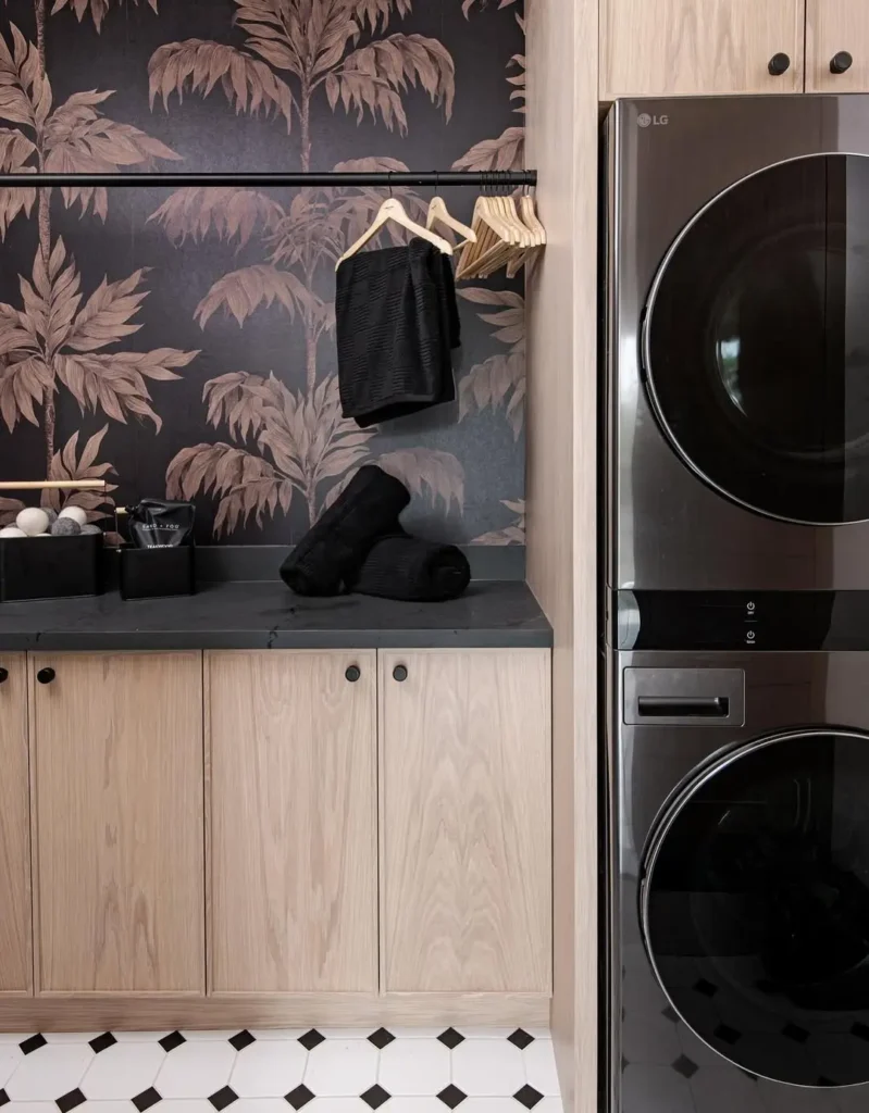 A modern laundry room with light wood cabinets, a black countertop, folded black towels, wooden hangers, stacked washer and dryer, and striking moody wallpaper with palm leaf designs.