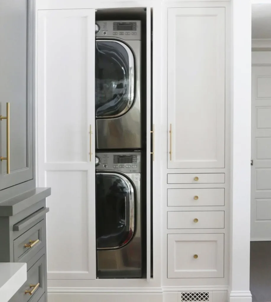 A stacked washer and dryer set is built into a tall white cabinet with gold handles, partially concealed by a sliding door—one of many smart ideas to fix common laundry room design mistakes.