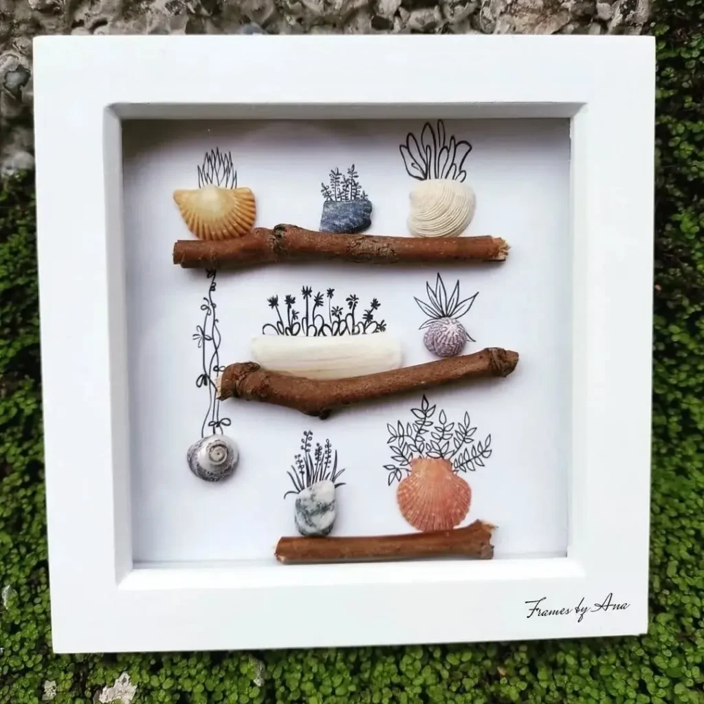 A white shadow box frame displays seashells, stones, and twigs arranged on drawn botanical designs against a textured mossy surface—a unique take on seashell wall art ideas.