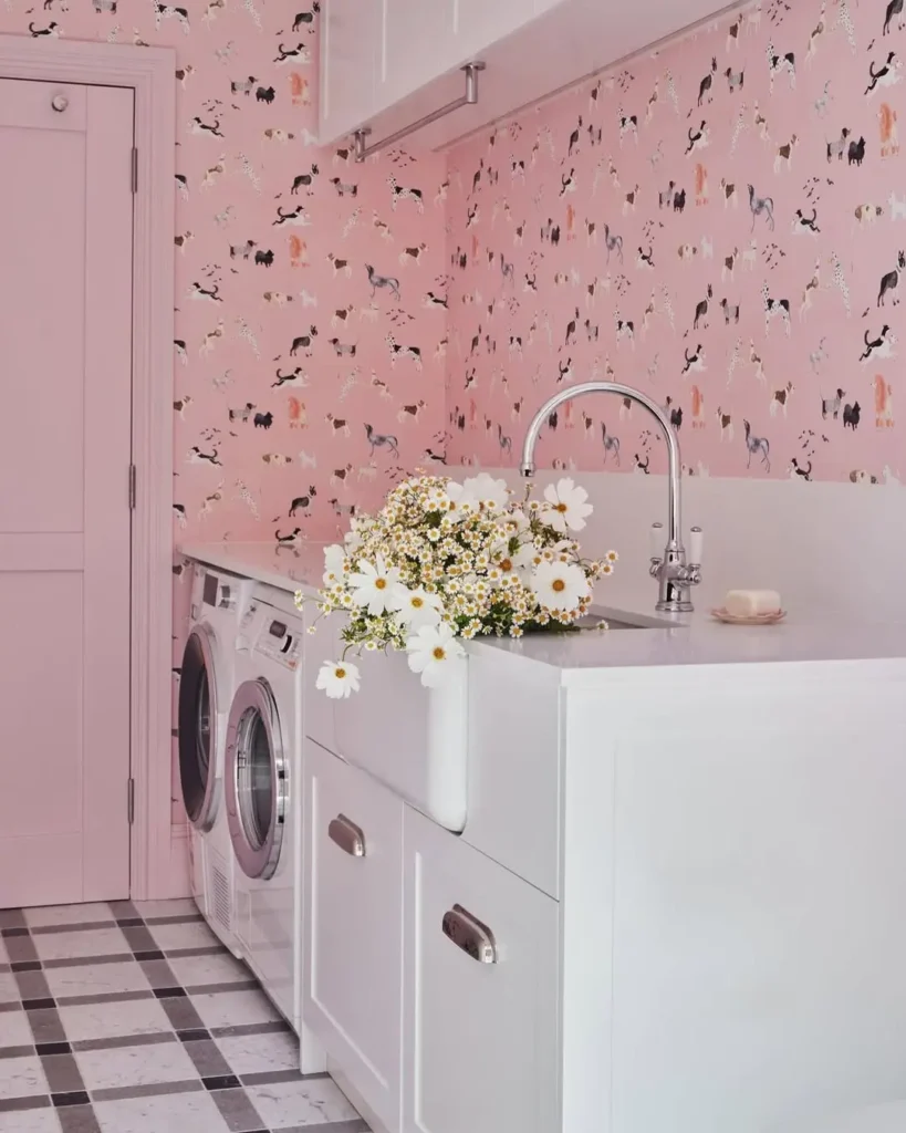A pink laundry room with playful pink wallpaper, a white sink and cabinets, front-loading washer and dryer, and a bouquet of daisies on the counter.