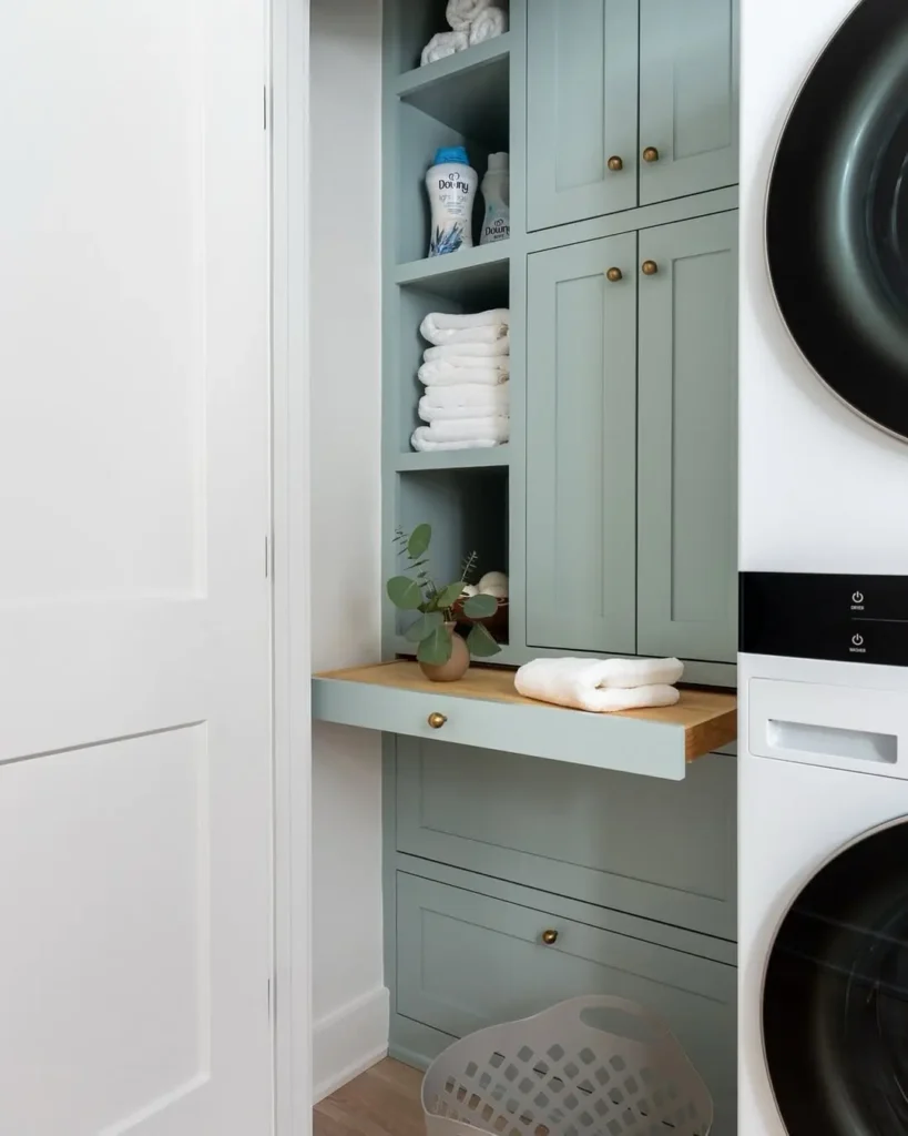 A compact laundry area with pale green cabinets, neatly stacked towels, detergent, and a small plant on a pull-out shelf—plus added counter space with a pull out folding station to avoid a common laundry mistake. White basket beside the front-load washer and dryer.