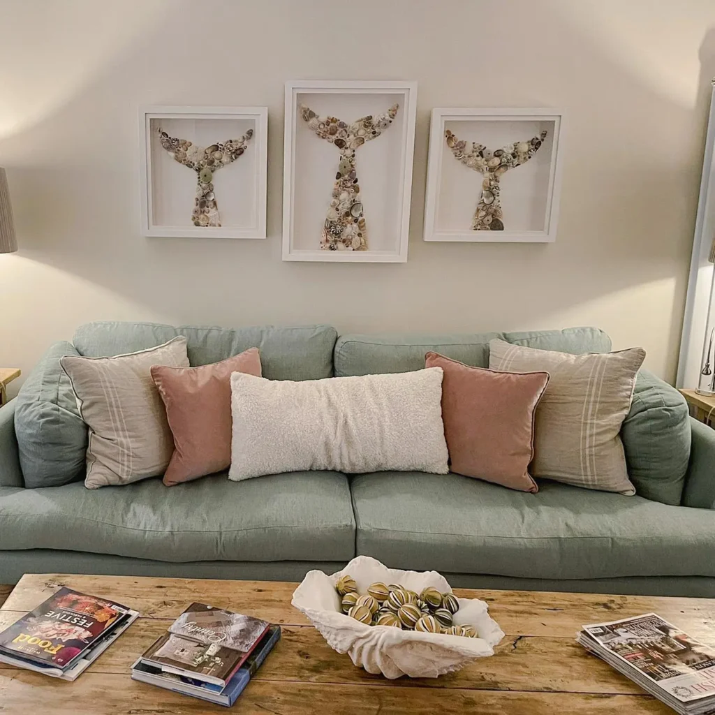 A light green couch with pink and white pillows sits against a wall with three framed whale tail artworks above. A wooden coffee table holds magazines and a bowl of decorative items.