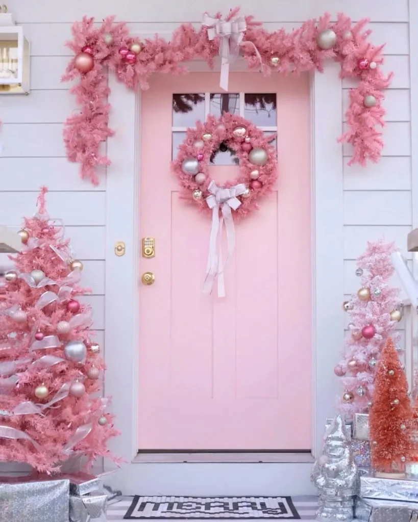 A pastel pink front door decorated with a pink wreath, garland, and matching pink Christmas trees, shatterproof Christmas ornaments, baubles, and wrapped gifts.