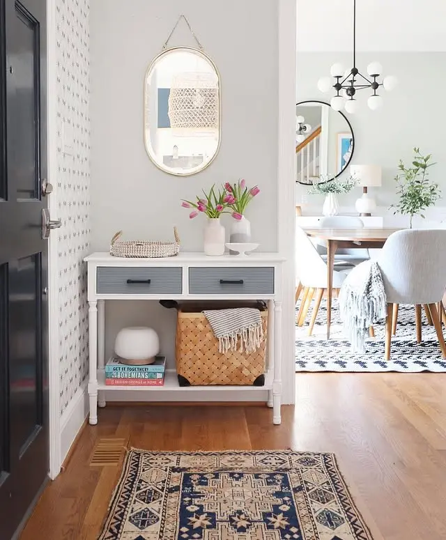 A stylish small entryway features a black door, white table with drawers, decorative items, bold-patterned floor rug, and a round mirror above, offering a glimpse into the dining room with its modern chandelier.