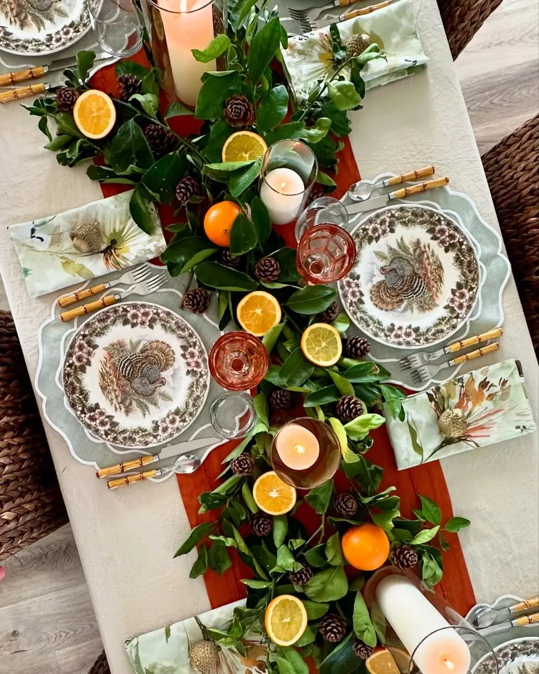 A Thanksgiving table decor scene featuring turkey-themed plates, floral napkins, gold utensils, and a centerpiece with greenery, pinecones, candles, and sliced citrus fruits.