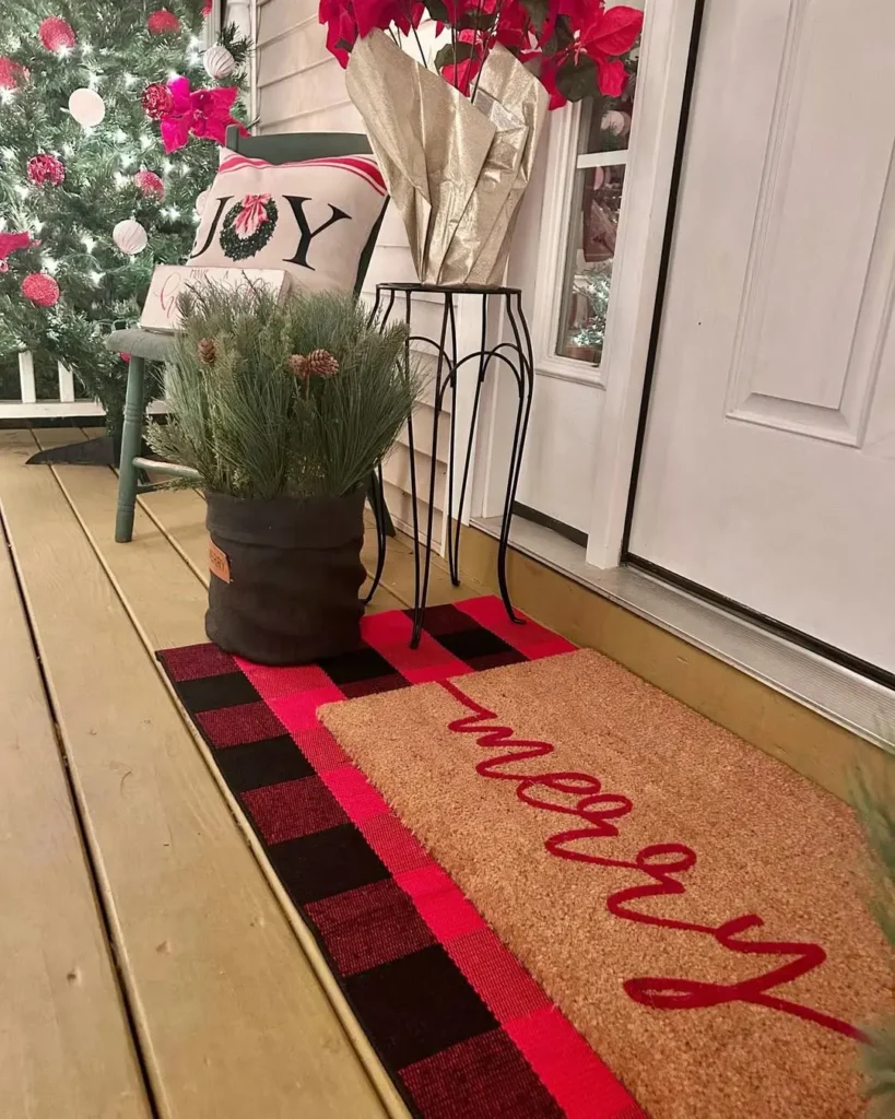 A holiday-themed winter porch doormat sets the scene alongside a "merry" mat, plaid rug, potted greenery, a "JOY" pillow, poinsettias, and part of a decorated Christmas tree.
