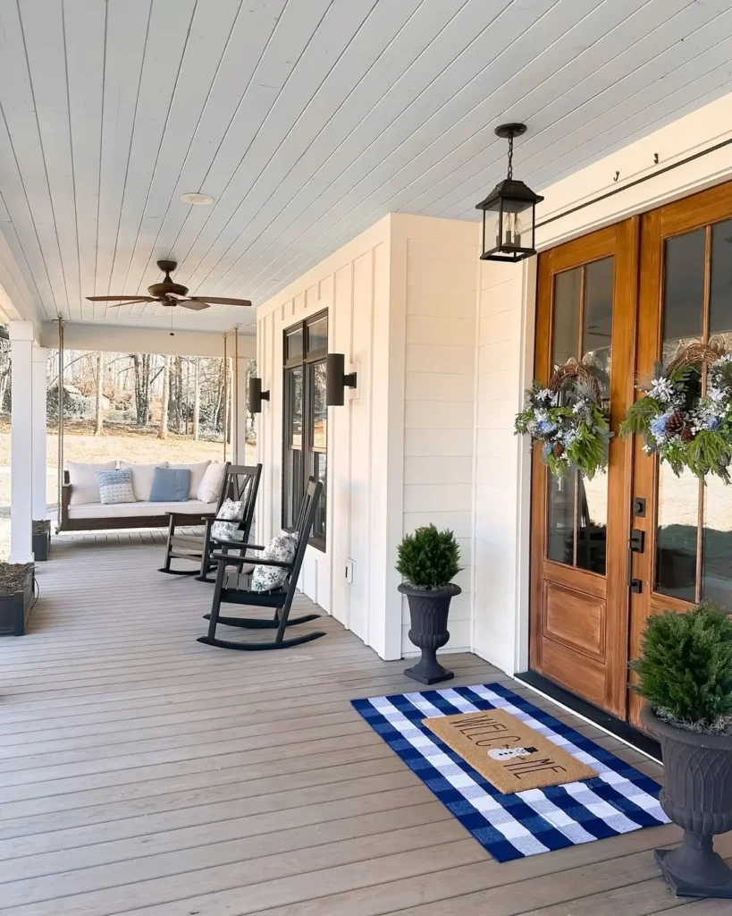 A farmhouse-style porch with rocking chairs, a cushioned swing, potted plants, and double doors decorated with wreaths. A cozy winter doormat adds charm to the blue and white rug by the entrance, completing this inviting porch decor.
