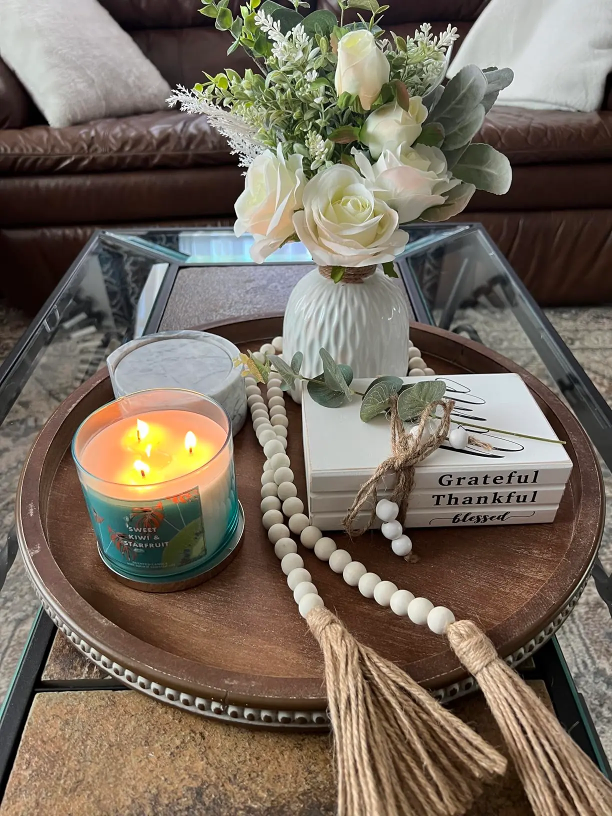 A round wooden tray with a lit candle, a white vase of artificial flowers, a beaded tassel garland, and stacked "Grateful Thankful Blessed" books creates charming Farmhouse Spring & Easter decor on a glass coffee table.