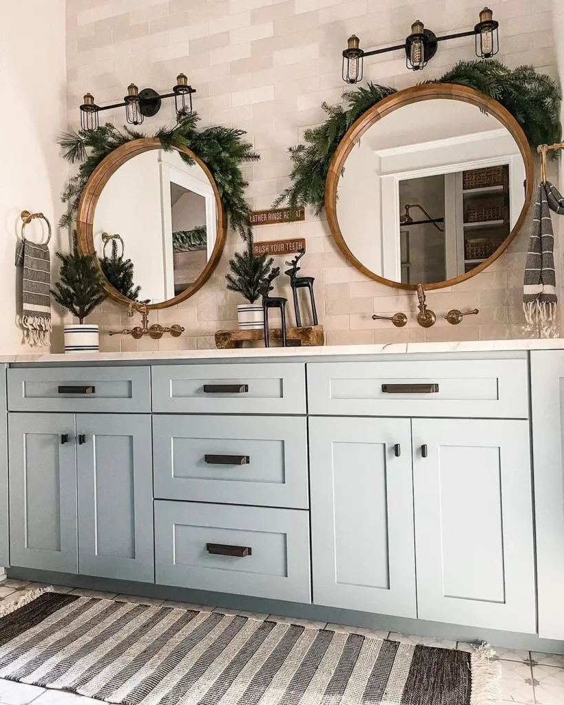 Double-sink bathroom vanity with light blue cabinets, round wood-framed mirrors, wall-mounted lights, and festive farmhouse bathroom Christmas decor with greenery above each sink.