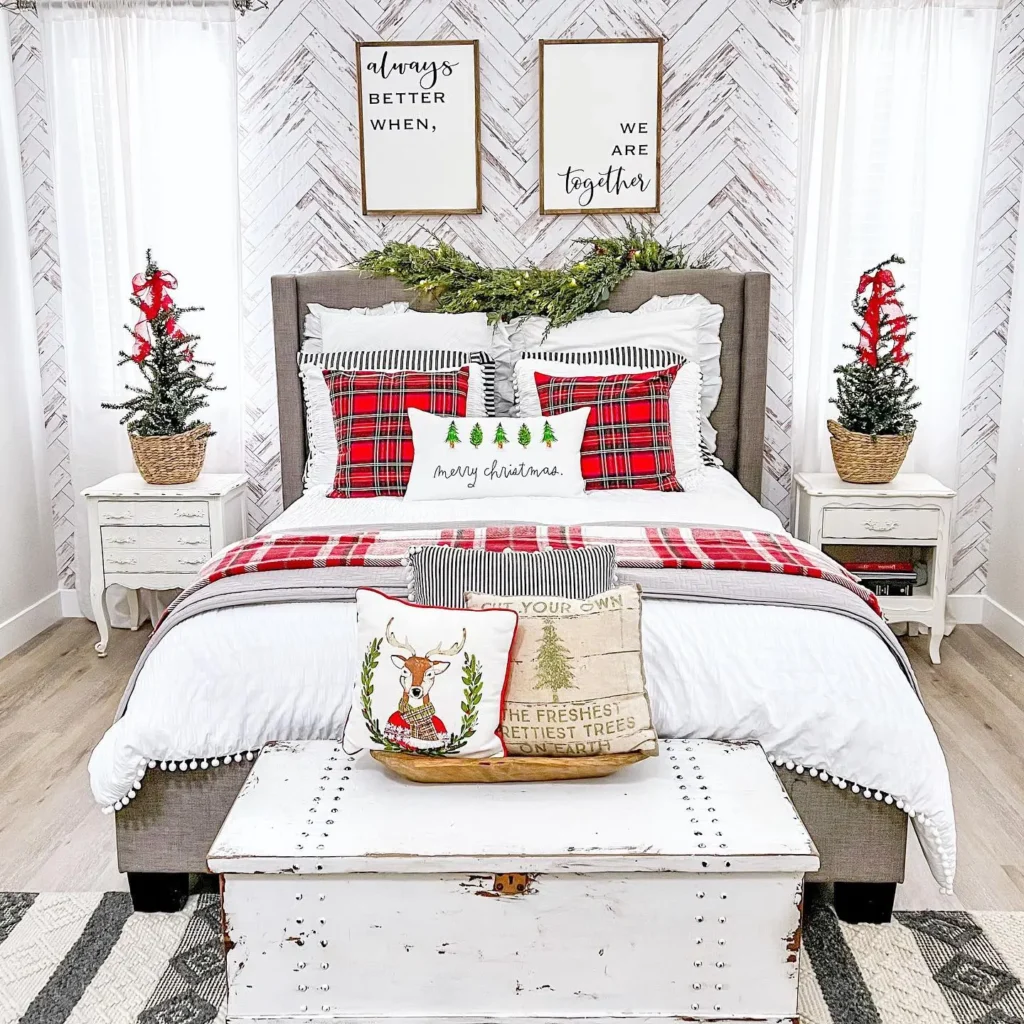 A neatly made bed with red plaid pillows, Christmas-themed cushions, small Christmas trees on nightstands, and framed quotes on a white paneled farmhouse wall.