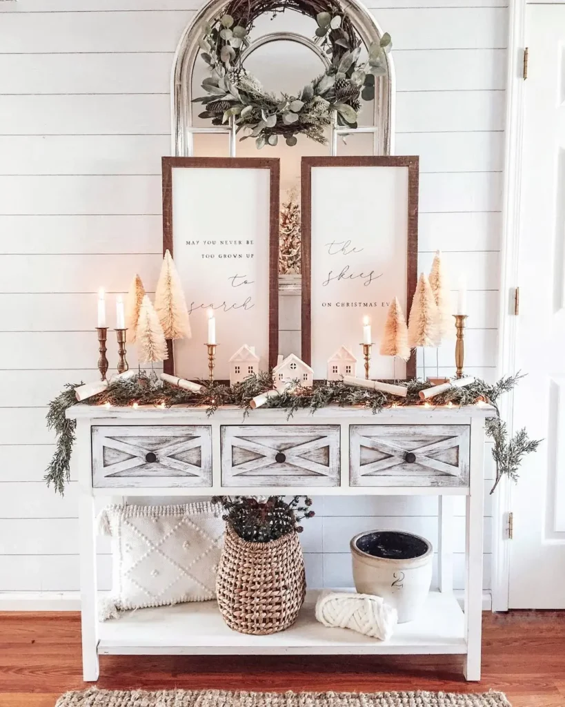A white console table styled with rustic farmhouse entryway Christmas decor, framed quotes, mini trees, candles, garland, and small houses completes the look, while baskets and pillows on the lower shelf add cozy charm.