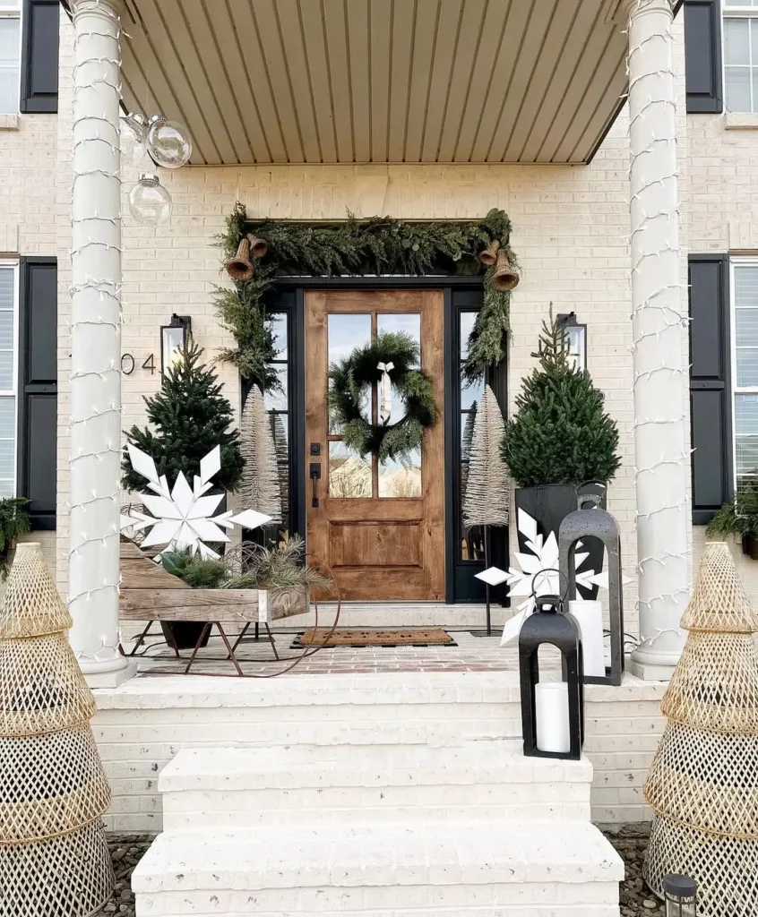 A front porch decorated for Christmas with modern rustic garlands, a wreath on the door, potted evergreen trees, white star ornaments, and lanterns with candles—all blending cozy farmhouse decor charm.