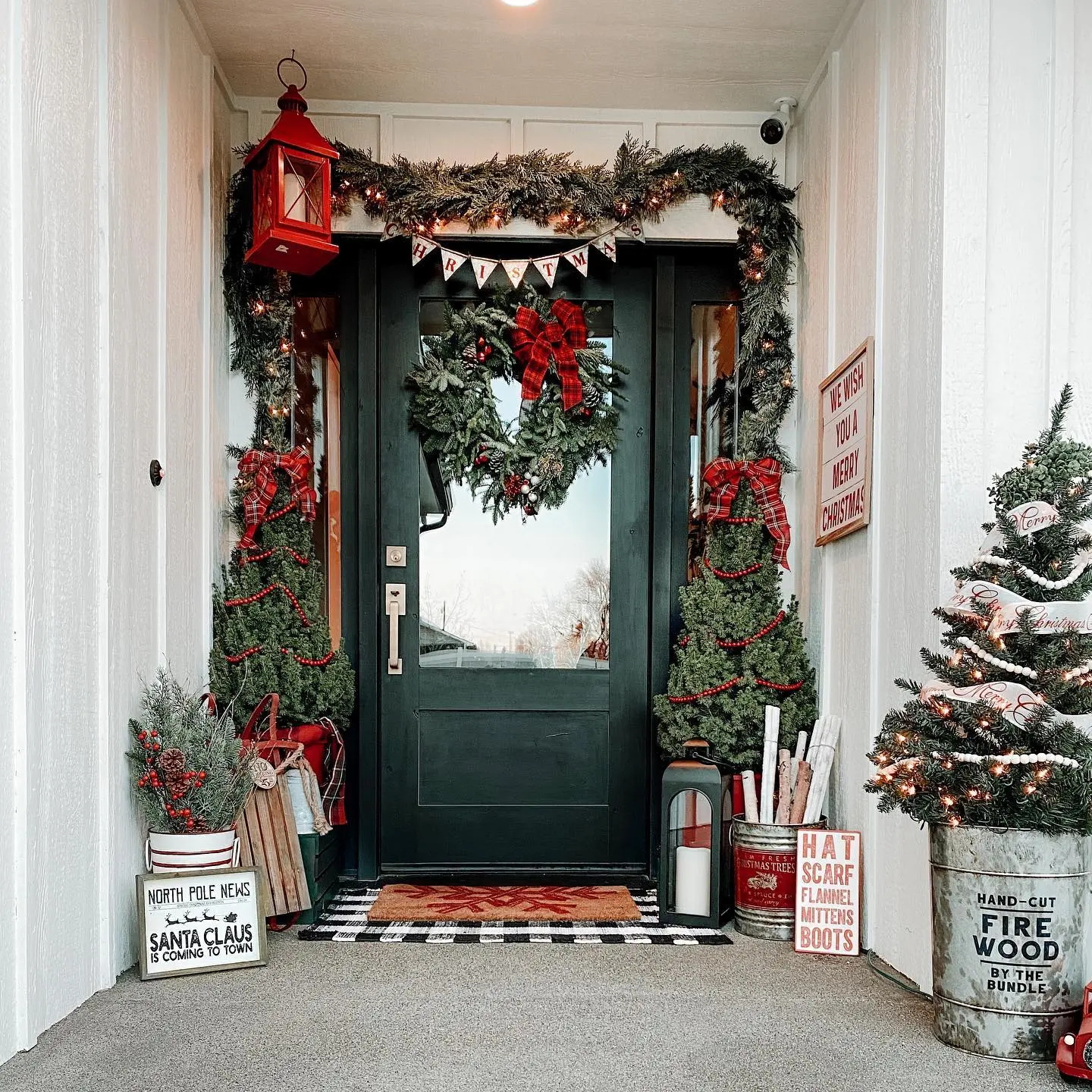 A front porch decorated for Christmas with a wreath, garlands, mini trees, lantern, and various holiday signs and ornaments.