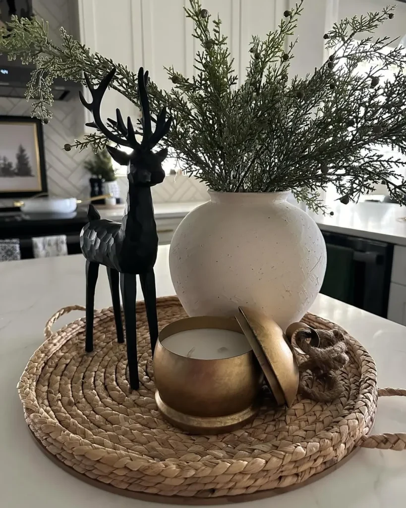 A decorative tray on a kitchen counter brings a touch of Christmas charm, featuring a black deer figurine, a white vase with green foliage, a gold candle with lid, and a knotted ornament.