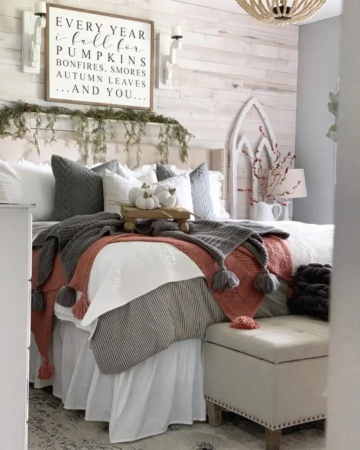 A cozy, well-decorated bedroom features layered bedding, autumn-themed pillows, a pumpkin centerpiece, and farmhouse fall bedroom decor, including a wall sign with a fall quote above the headboard.