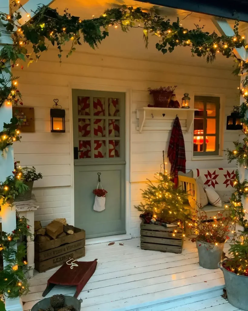 A small porch decorated with string lights, greenery, a mini Christmas tree in a crate, a bench with red pillows, and a stocking hanging on the front door.
