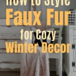 A cozy winter living space featuring a soft blush faux fur throw draped over a white chair, with rustic farmhouse-style furniture, a tulip-filled wall basket, and warm wood flooring. Text overlay reads: “How to Style Faux Fur for Cozy Winter Decor.”