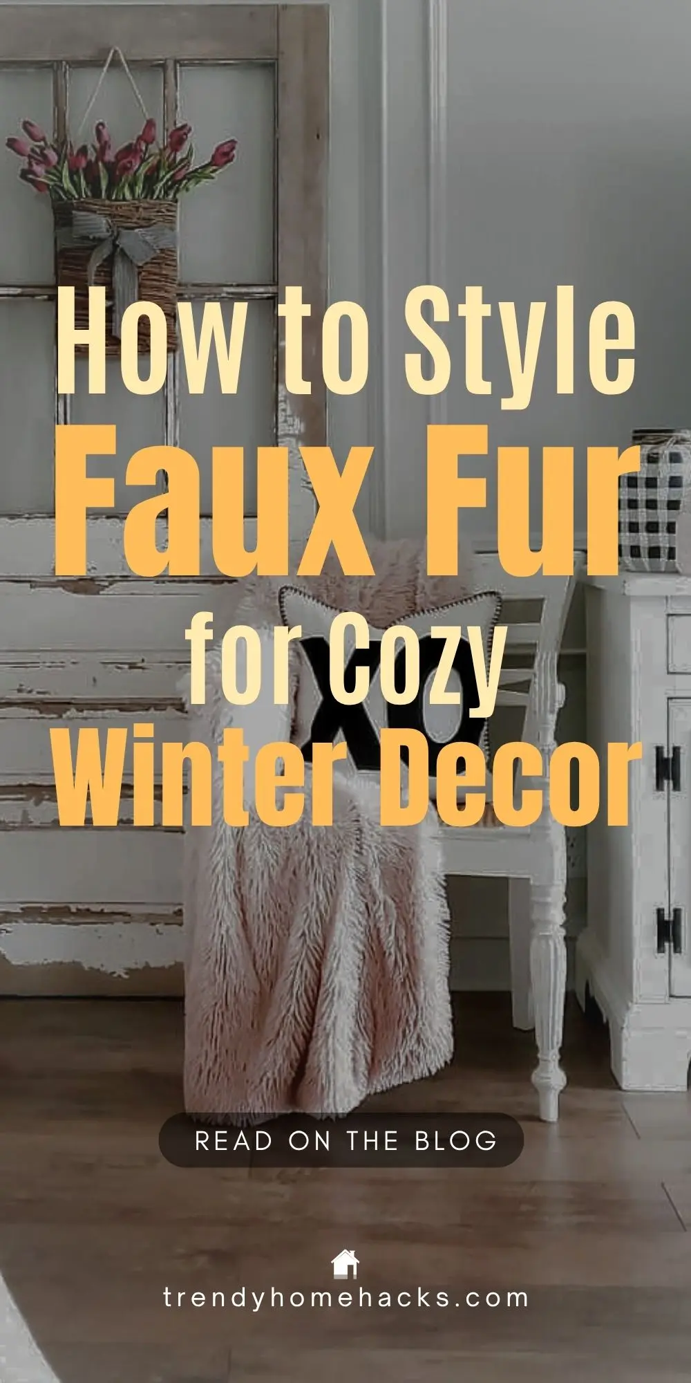 A cozy winter living space featuring a soft blush faux fur throw draped over a white chair, with rustic farmhouse-style furniture, a tulip-filled wall basket, and warm wood flooring. Text overlay reads: “How to Style Faux Fur for Cozy Winter Decor.”
