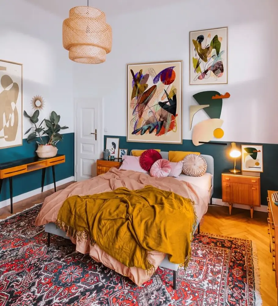 A colorful bedroom features maximalist fall layering tips with a double bed in mustard bedding, decorative pillows, abstract wall art, wooden furniture, a red patterned rug, and a woven ceiling light.