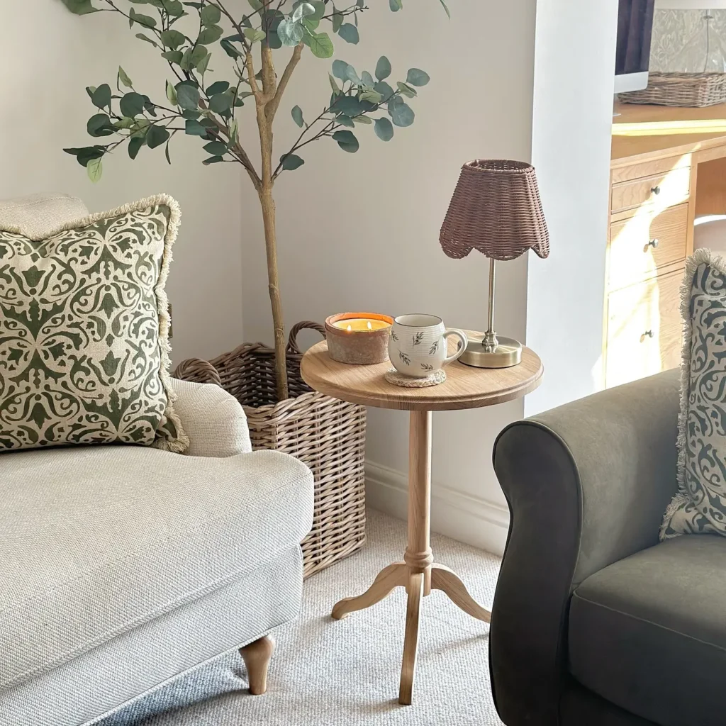 A small round wooden side table in a compact living space, holds a mug, candle, and lamp between a beige sofa and gray armchair, with a potted plant and wicker basket in the background.