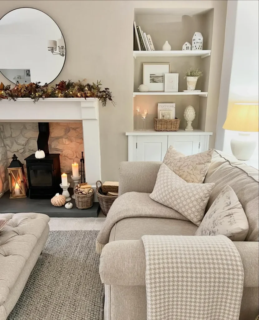 A cozy living room with a beige sofa, patterned cushions, a fireplace adorned with thanksgiving decor with candles and string lights, built-in shelves, and a round mirror above the mantel decorated with foliage.