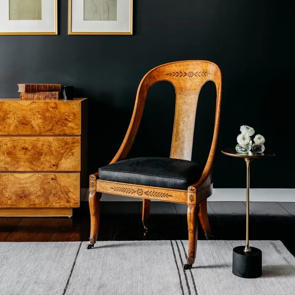 A bedroom with a rustic wood and leather accent chair that sits between a wood dresser and a small round side table with white flowers, set against a dark wall adorned with framed art.