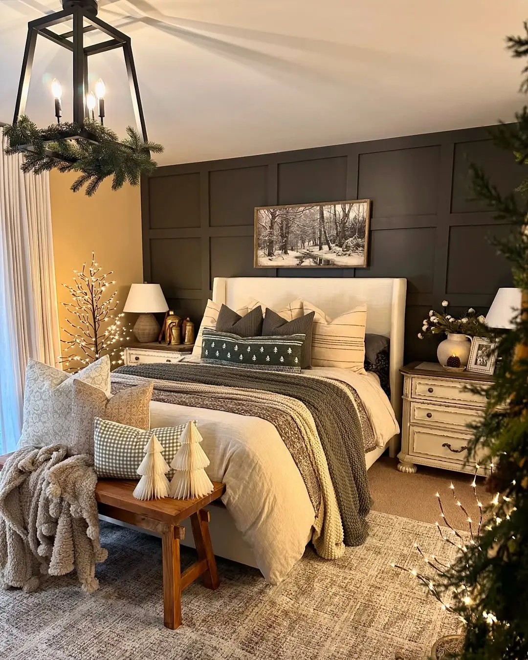 A cozy bedroom features a bed with layered pillows and blankets, a wood bench with tree decorations, bedside tables with lamps, a winter landscape photo on the wall, and festive holiday accents.