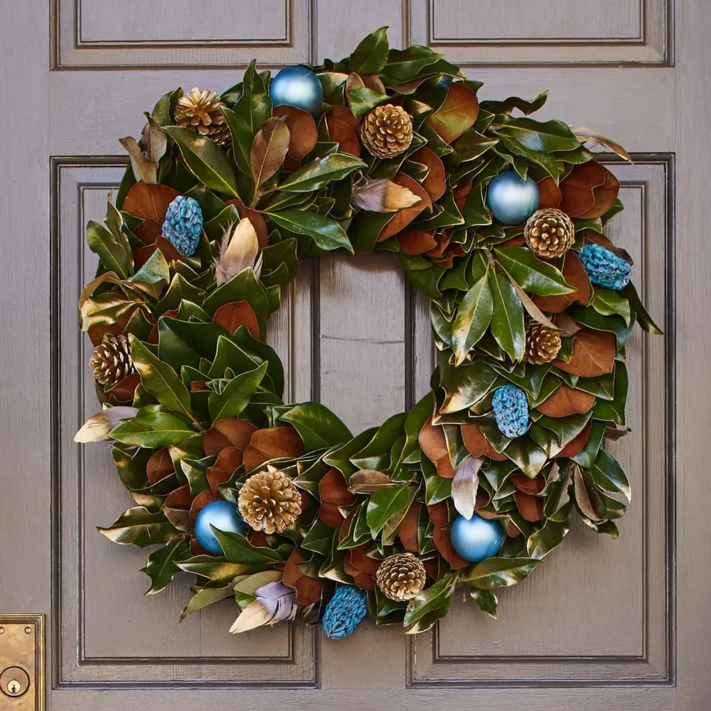 A winter wreath made of green and brown leaves, pinecones, and blue ornaments hangs on a gray paneled door.