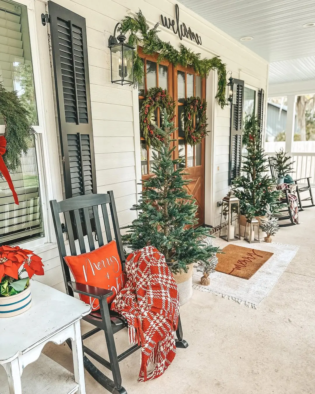 Christmas front porch decor ideas featuring green rocking chairs with red plaid throws, small potted Christmas trees, and a double wreath front door framed with garland for a cozy country farmhouse look.