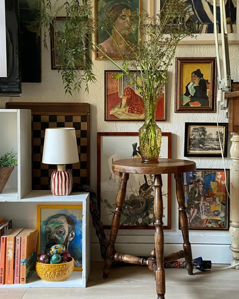A wooden table with a green vase of wildflowers stands before a maximalist gallery wall filled with framed artworks; a lamp and books sit on nearby shelves.
