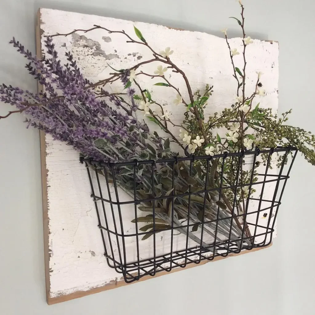 A black wire basket mounted on a distressed white wooden board brings farmhouse charm as it holds artificial lavender, greenery, and small white flowers against a light gray wall.