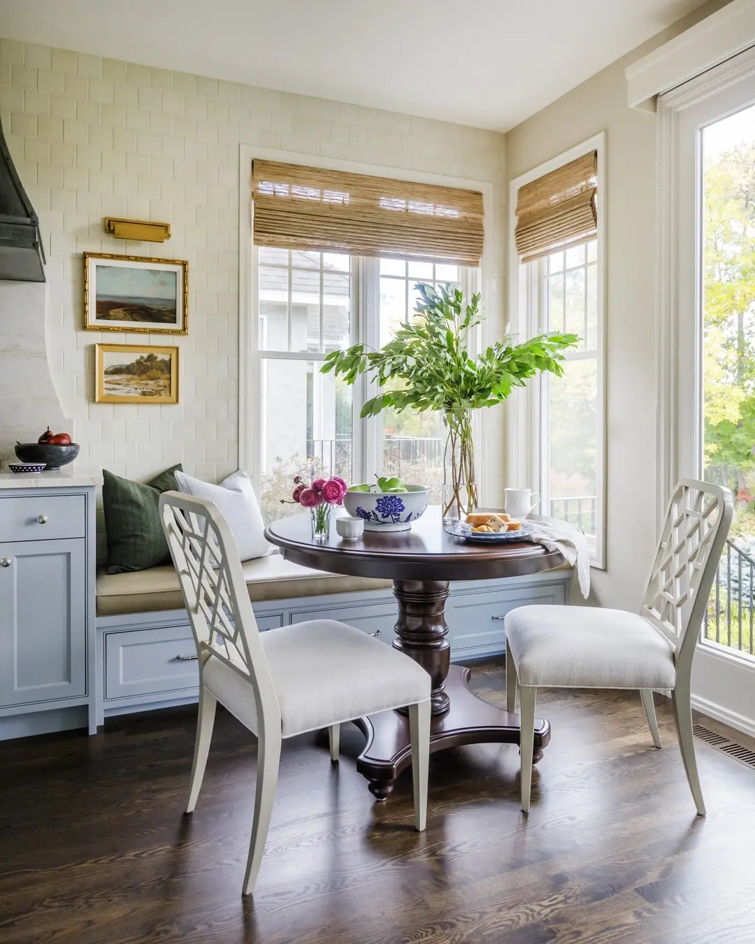 A round wooden table with two cushioned chairs sits by large windows; a vase with green foliage and plates of food are on the table. Built-in banquette seating with drawers and wall art are visible in the background.